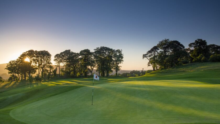A golf green with a flagstick is shown at sunset, with long shadows from nearby trees and a clear sky in the background.