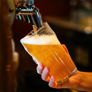 A hand holds a glass at an angle under a beer tap as golden beer with a frothy head is poured.