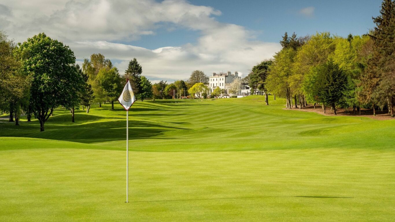 A well-maintained golf course with a flag marking a hole in the foreground, surrounded by trees, and a large white building in the distance under a partly cloudy sky.