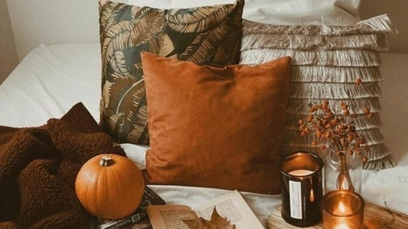 A cozy bed with pillows, a brown blanket, a pumpkin, candles, a cup of coffee, an open book, and fall leaves arranged on a white bedsheet.
