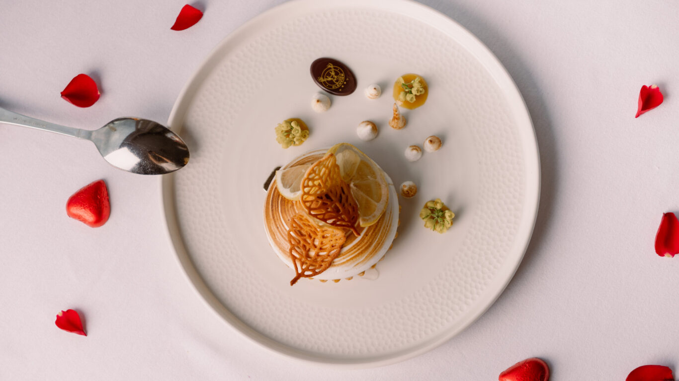 Elegant dessert with decorative elements on white plate, surrounded by red rose petals on table, offering a romantic dining experience.