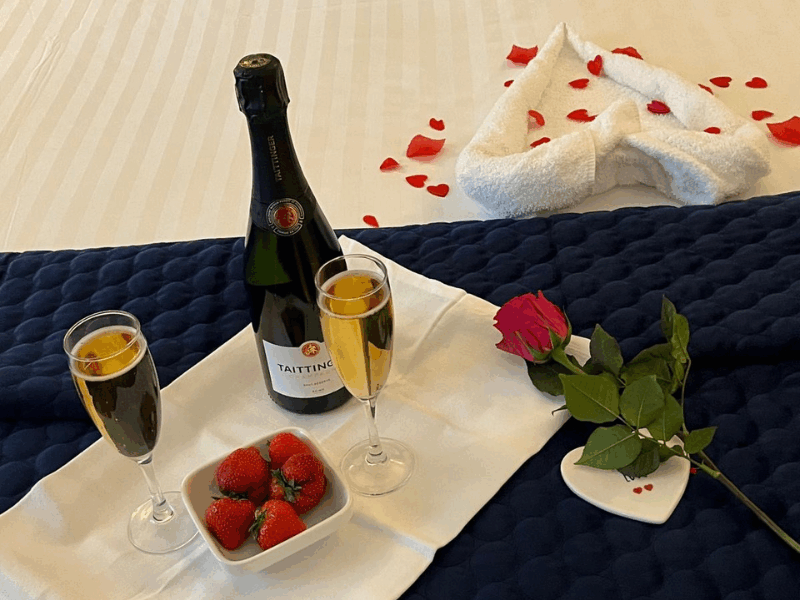 A bottle of champagne with two filled glasses, a bowl of strawberries, and a single rose on a bed with rose petals and towel art in the background.