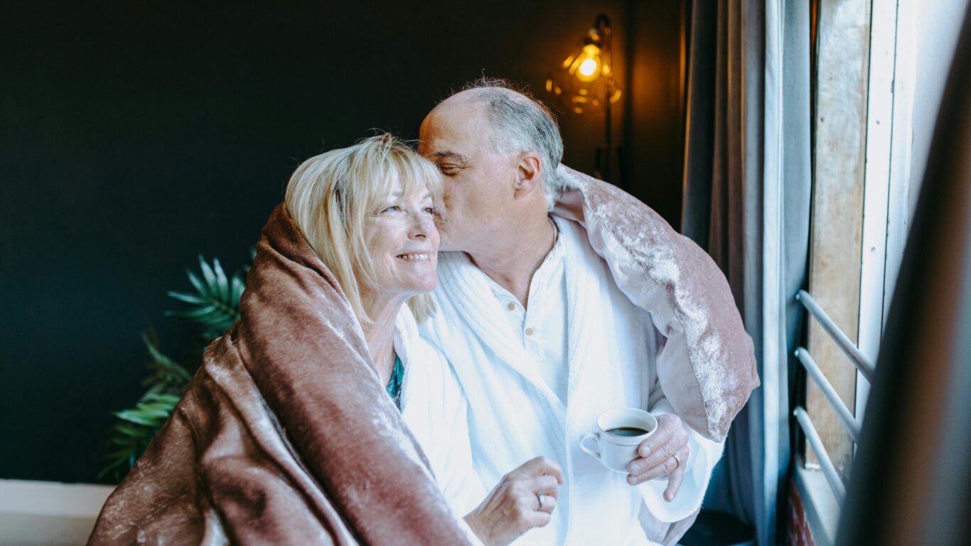 Mature couple enjoys cosy morning with coffee by hotel window, wrapped in soft blankets, basking in warm ambient lighting.