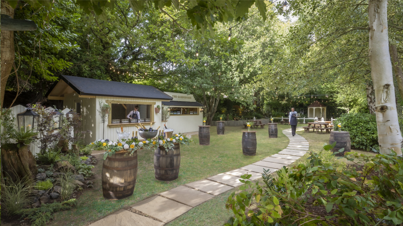 Picturesque garden bar with wooden benches under lush trees, offering an inviting outdoor space for relaxation and socialising.