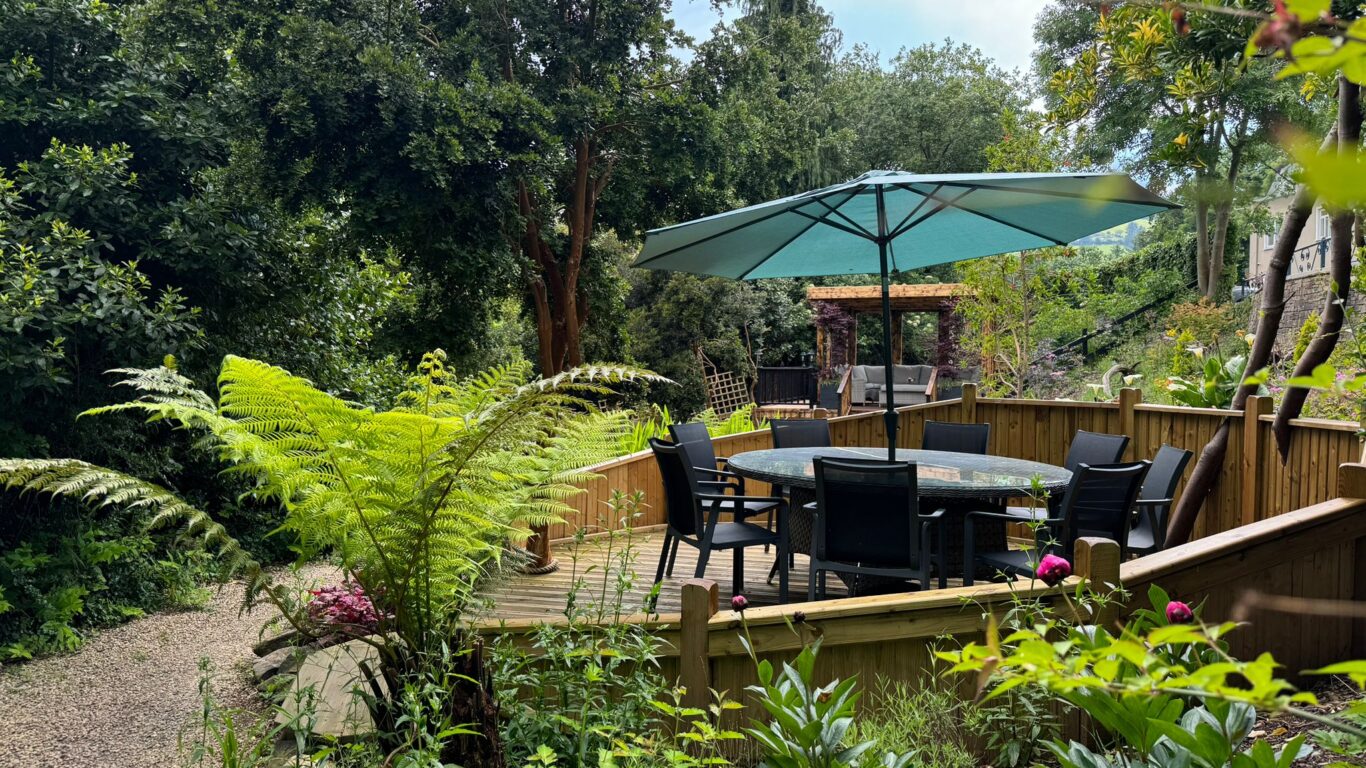 A round table with six chairs and a large green umbrella sits on a wooden deck surrounded by lush greenery and a gravel path in a garden.