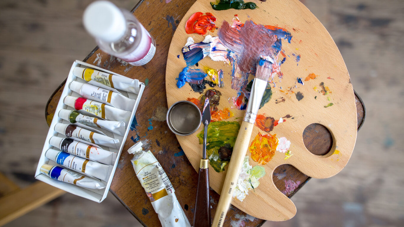 An artist's palette with mixed paints, paintbrushes, a box of paint tubes, and a bottle of liquid, all placed on a wooden surface.