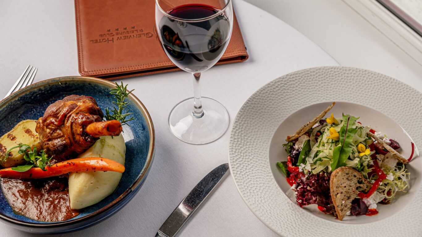 A plate with lamb shank, mashed potatoes, and vegetables next to a salad, a glass of red wine, and a closed menu on a white tablecloth.