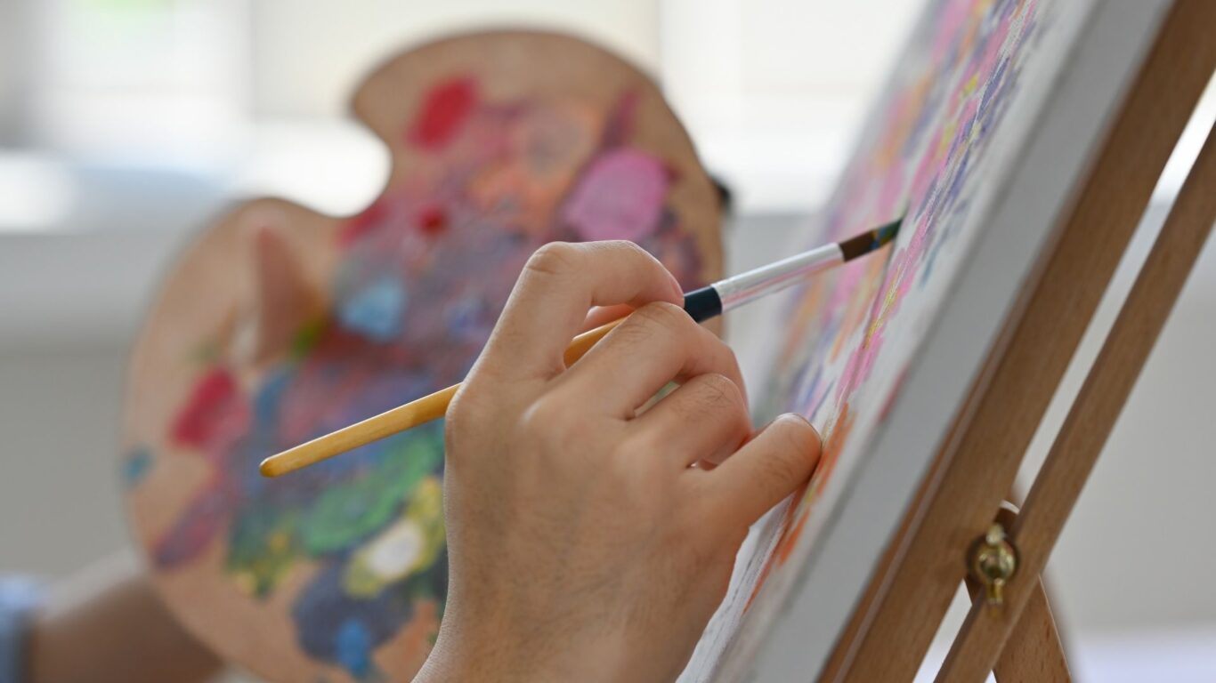 A person holds a paintbrush and palette while painting on a canvas mounted on an easel.