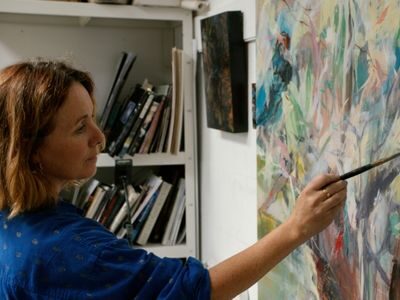 A woman in a blue shirt paints on a colorful canvas in a studio, with shelves of books and art supplies in the background.