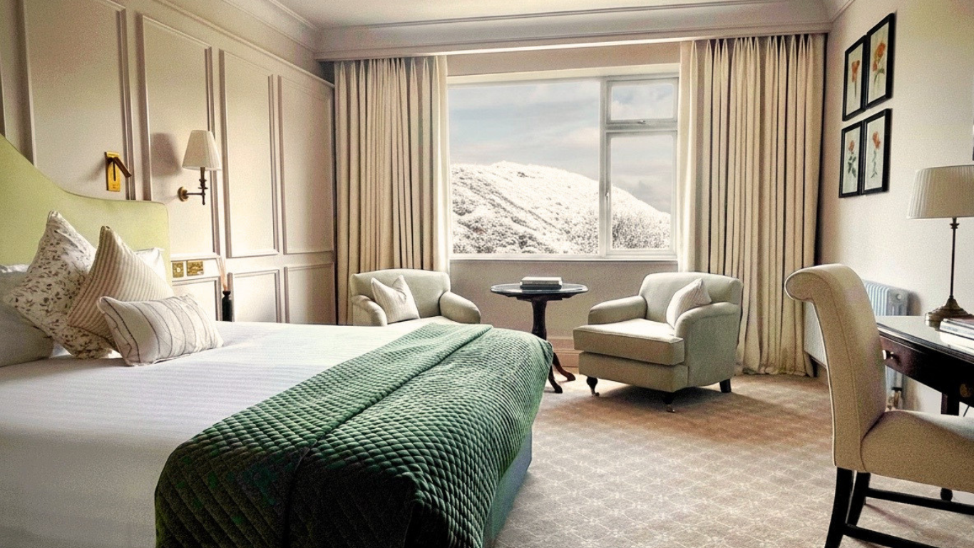 Spacious hotel room with plush bed, armchairs, scenic mountain view through large window, inviting relaxation.